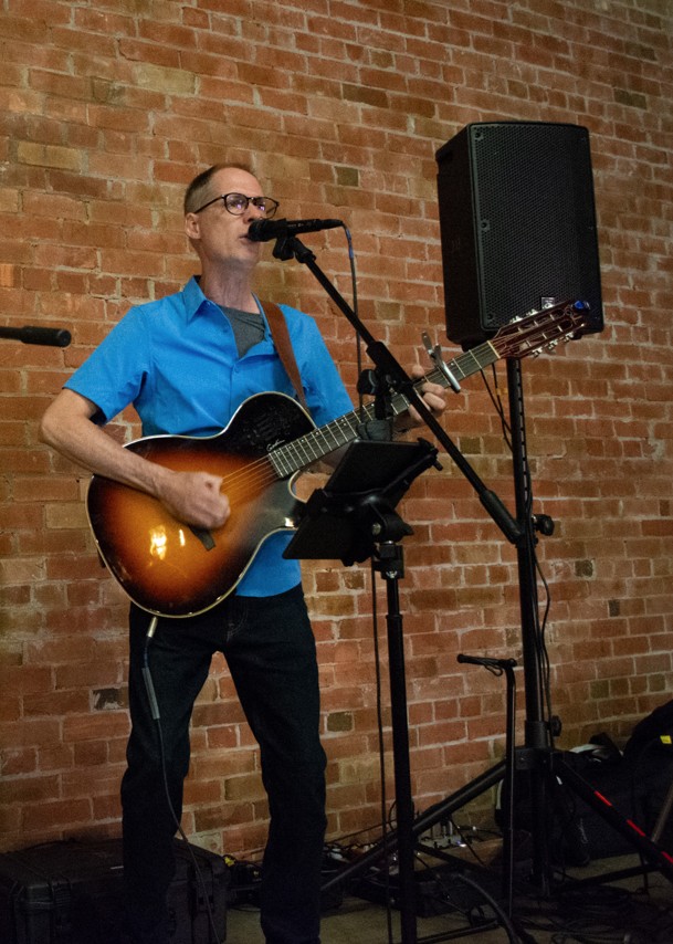 Jon performing guitar and vocals in front of a brick wall